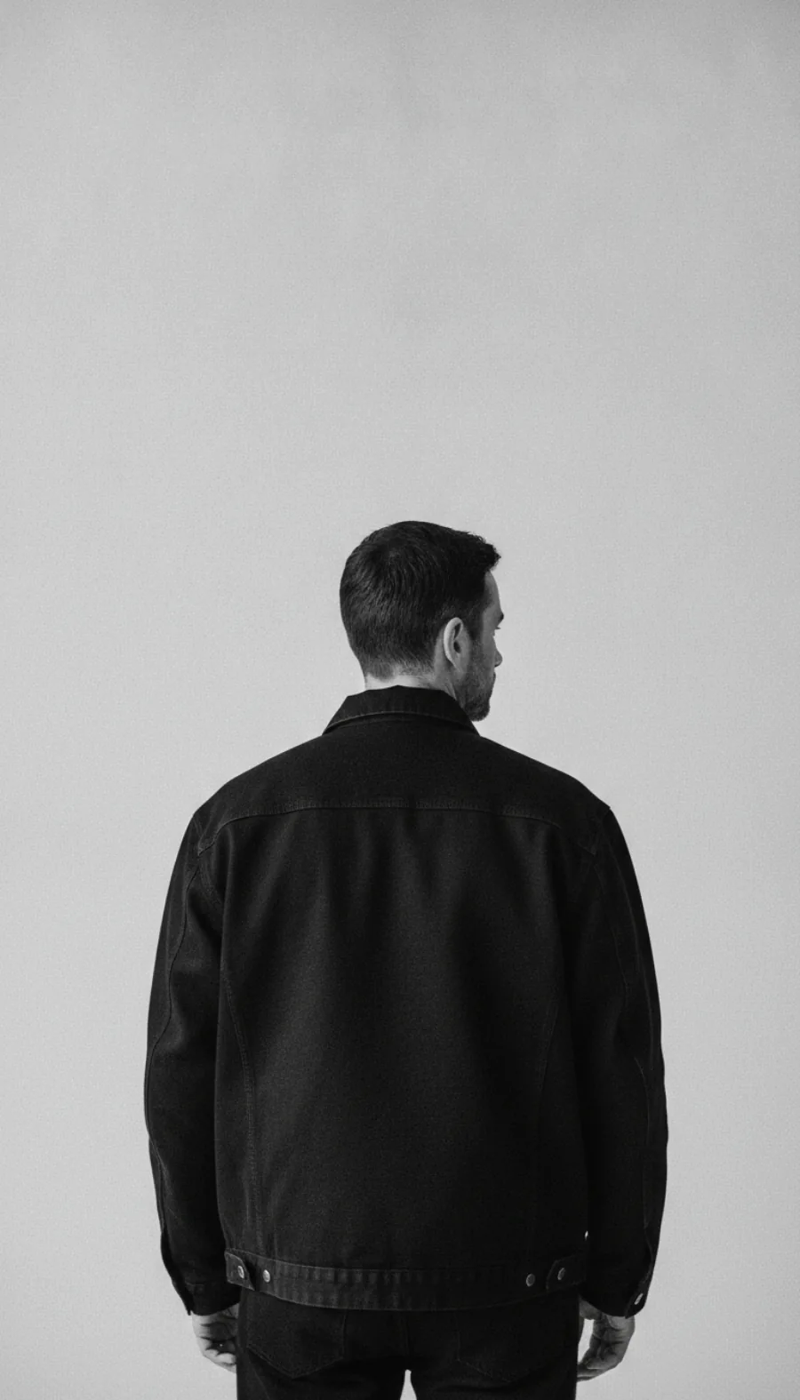 Un homme de dos, en veste de travail noire, devant un fond gris